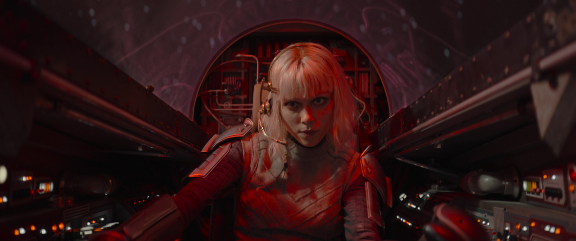 Shin Hati (Ivanna Sakhno), a human with blonde hair and blunt bangs, sits in the cockpit of a spaceship, her expression focused as she pilots it. She is wearing a headset with a microphone and is bathed in red light.