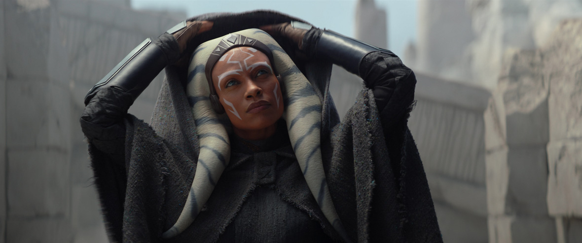 Ahsoka Tano (Rosario Dawson), an orange-skinned Togrutan with white face markings, lifts the hood of her cloak off of her head with a serious expression.