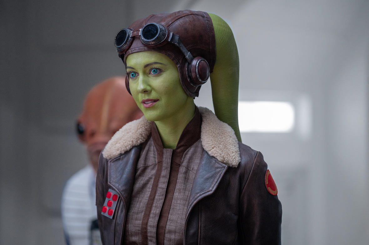 Hera Syndulla (Mary Elizabeth Winstead), a green Twi’Lek wearing a flight jacket and goggles, looks at something offscreen to the left.