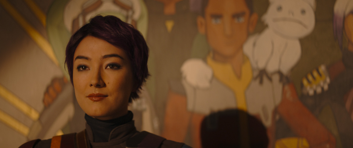 Sabine Wren (Natasha Liu Bordizzo), a Mandalorian with short, purple hair, stands in front of a mural she painted featuring the crew of the Ghost. She is looking off into the distance with a thoughtful expression.