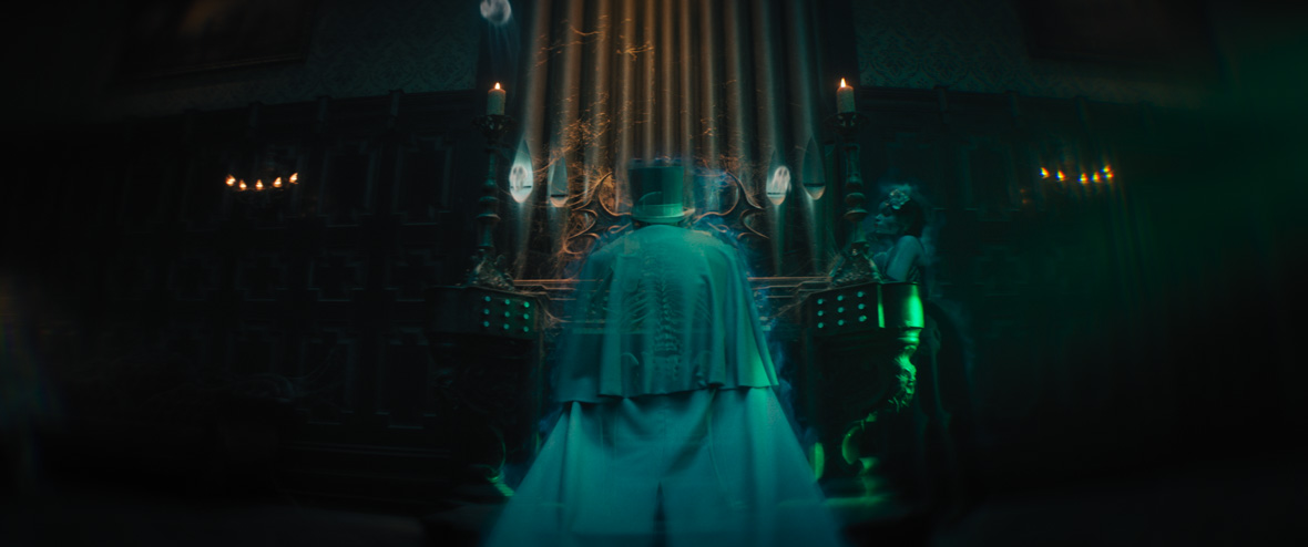 A teal, transparent ghost in a top hat and cape has his back to us as he plays on a pipe organ. Most of the room is in darkness, except for the ghost, his instrument, and a ghostly woman leaning on the right side of the organ.