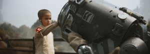 In a scene from the film The Creator, Alphie, played by Madeleine Yuna Voyles, is wearing a beige long-sleeve shirt with a red patch on her right shoulder while she puts her right hand on a metal canister-shaped robot that has 4 metal legs.