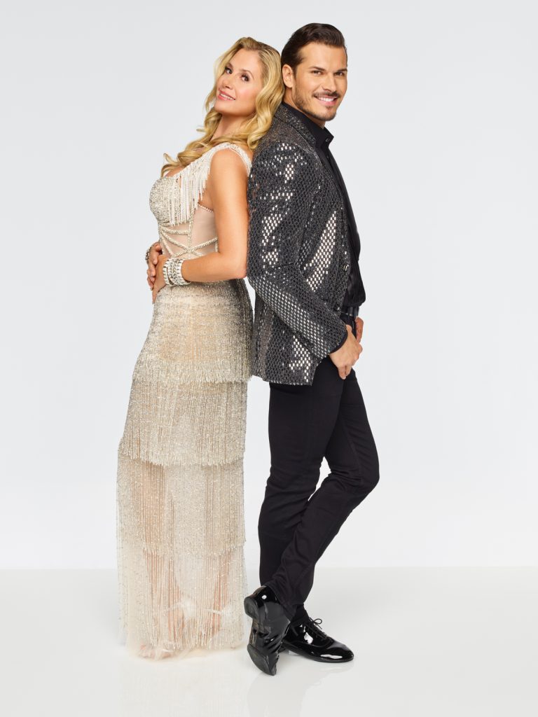 Mira Sorvino wears a floor length gold fringe tiered dress as she stands back to back with Gleb Savchenko who is wearing a dark silver reflective blazer with black pants.