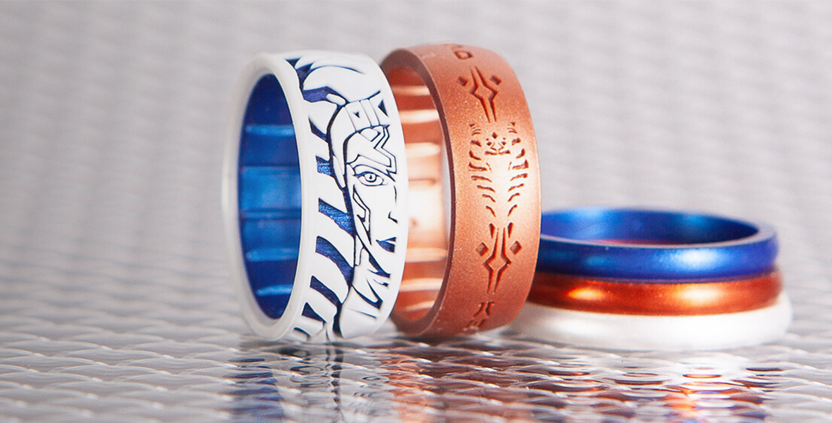 Three rings sit on a silver surface. One ring is laid on the surface, striped in orange, white, and blue. Two other rings are balanced on their sides next to each other. One ring is orange with Ahsoka Tano’s face marking inscribed on it. The other is blue and white with Ahsoka Tano’s face carved into it.