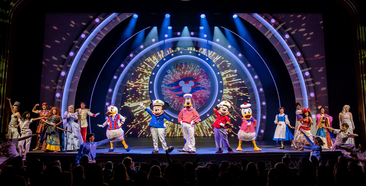 In a promotional image from Disney Cruise Line, the cast of Disney Seas the Adventure is seen on stage at the Walt Disney Theatre. Among the characters are Mickey Mouse, Goofy, Minnie Mouse, and Donald Duck in their nautical best, as well as Princess Tiana, Hercules, Merida, Cinderella, Belle, Aladdin, Moana, Mulan, and Elsa. Behind the cast, projected, is the Disney Cruise Line logo surrounded by virtual fireworks. Audience members can be seen in silhouette, applauding the show.