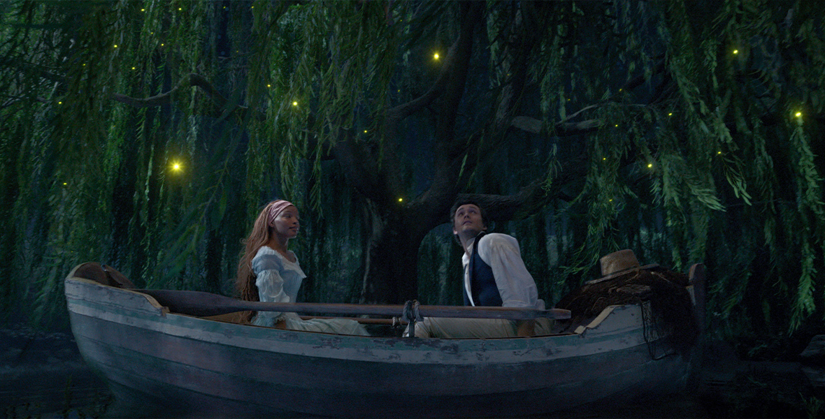 In an image from Disney’s live action The Little Mermaid, Ariel (Halle Bailey) and Prince Eric (Jonah Hauer-King) are sitting in a rowboat, surrounded by branches thick with swamp-like leaves; these branches and leaves are hanging down all around them, and are filled with fireflies, lending a romantic atmosphere.