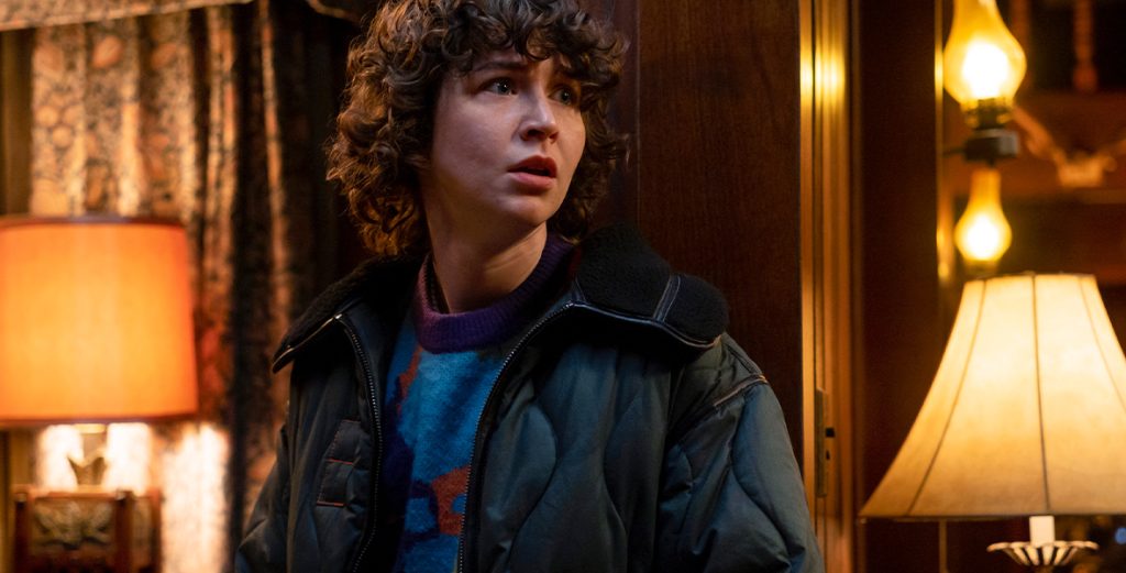 In the Goosebumps episode "Give Yourself Goosebumps," actor Miles McKenna appears frightened. McKenna is wearing a graphic sweater and a navy blue puffer jacket with orange stitching. The room features antique lamps, floral curtains, and wood paneling.