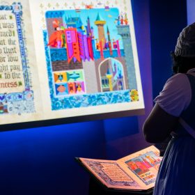 Two Disney fans look down at a replica of the prop book from Sleeping Beauty. In front of them is a larger image of the inside of their book, projected on a screen and backed by blue glowing light.