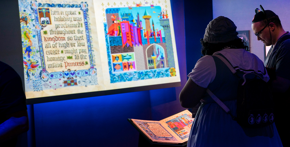 Two Disney fans look down at a replica of the prop book from Sleeping Beauty. In front of them is a larger image of the inside of their book, projected on a screen and backed by blue glowing light.