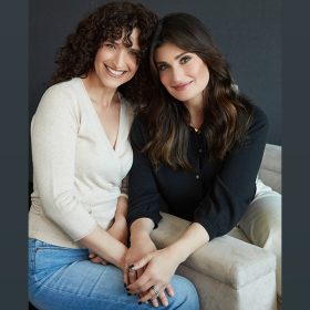 Cara Mentzel stands on the left, leaning toward her sister, Idina Menzel, who sits on a cream-colored sofa. Their heads are touching, and Idina has her hands resting atop her sister’s right hand. Cara’s dark hair is curly, and she wears a V-neck cream-colored shirt and light-colored jeans. She smiles fully at the camera. Idina has a closed-mouth smile, her dark hair blown out straight. She wears a black blouse, a small gold necklace with a heart visible, and white pants.