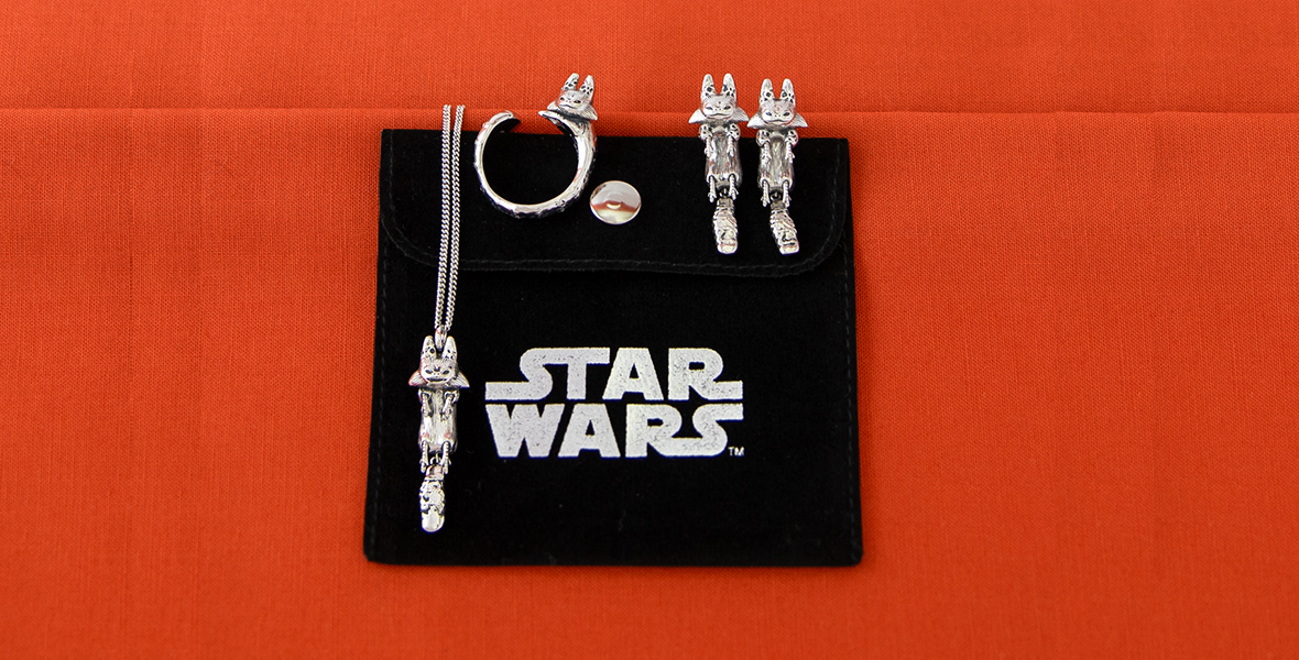 A black jewelery backer emblazoned with the Star Wars logo sits on a red oange background. On the jewelery back are silver earrings, a necklace, and ring, all shaped to look like the loth cat from the Ahsoka series.