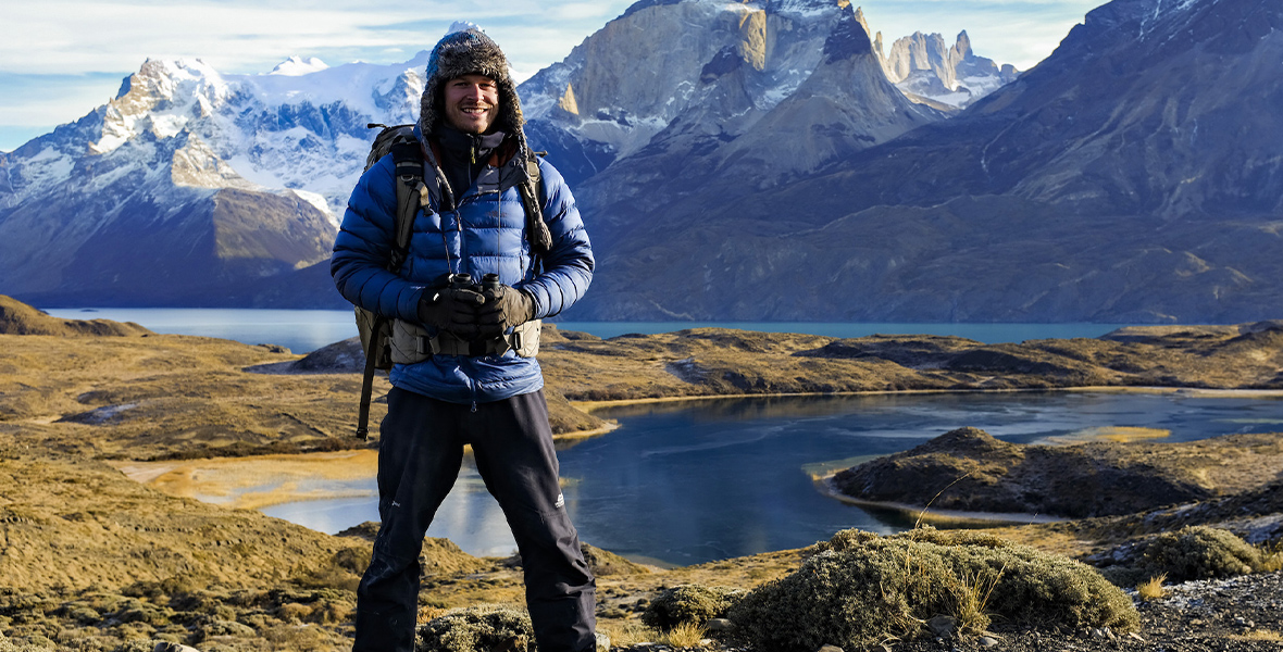 In an image from Animals Up Close with Bertie Gregory, the explorer is seen in Patagonia, with beautiful hills and snow-capped mountains rising behind him. There is also a river winding behind him, at the foot of the mountains. He is smiling at the camera, and is wearing a blue puffy jacket, dark blue pants, and a fur lined hat; he’s holding binoculars and is wearing a backpack.