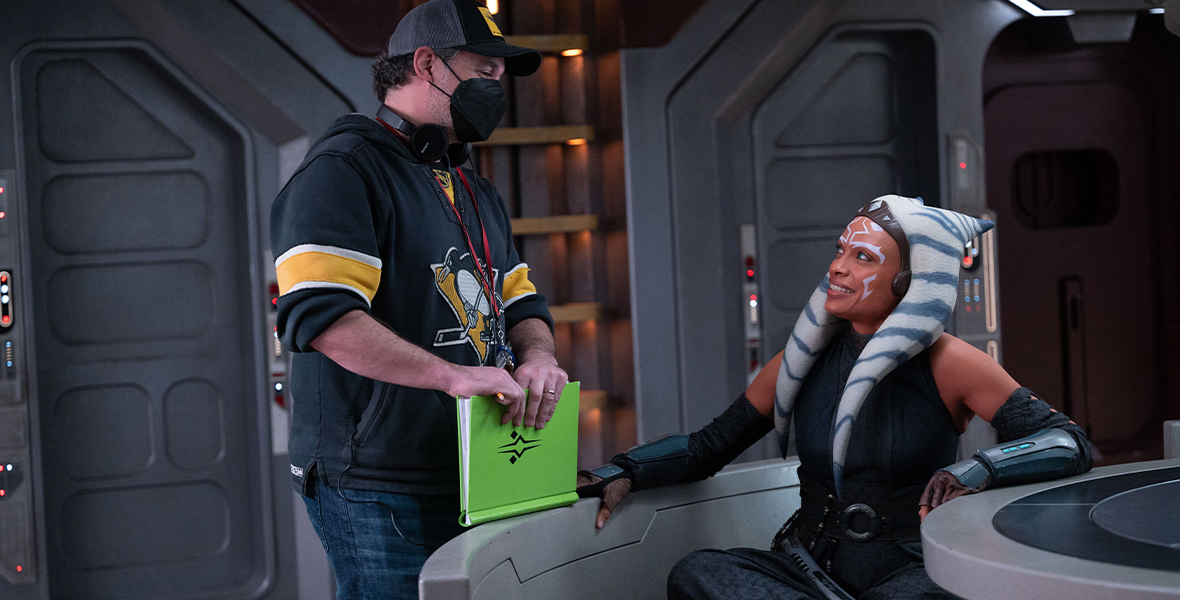 In a behind-the-scenes image from Lucasfilm’s Ahsoka, Disney Legend Jon Favreau (left) is seen—wearing a hoodie, jeans, a baseball cap, and a mask—standing next to Rosario Dawson as Ahsoka, who is seated. They’re on one of the sets for the series. Dawson is looking up at Favreau and smiling; he’s holding a bright green folder.