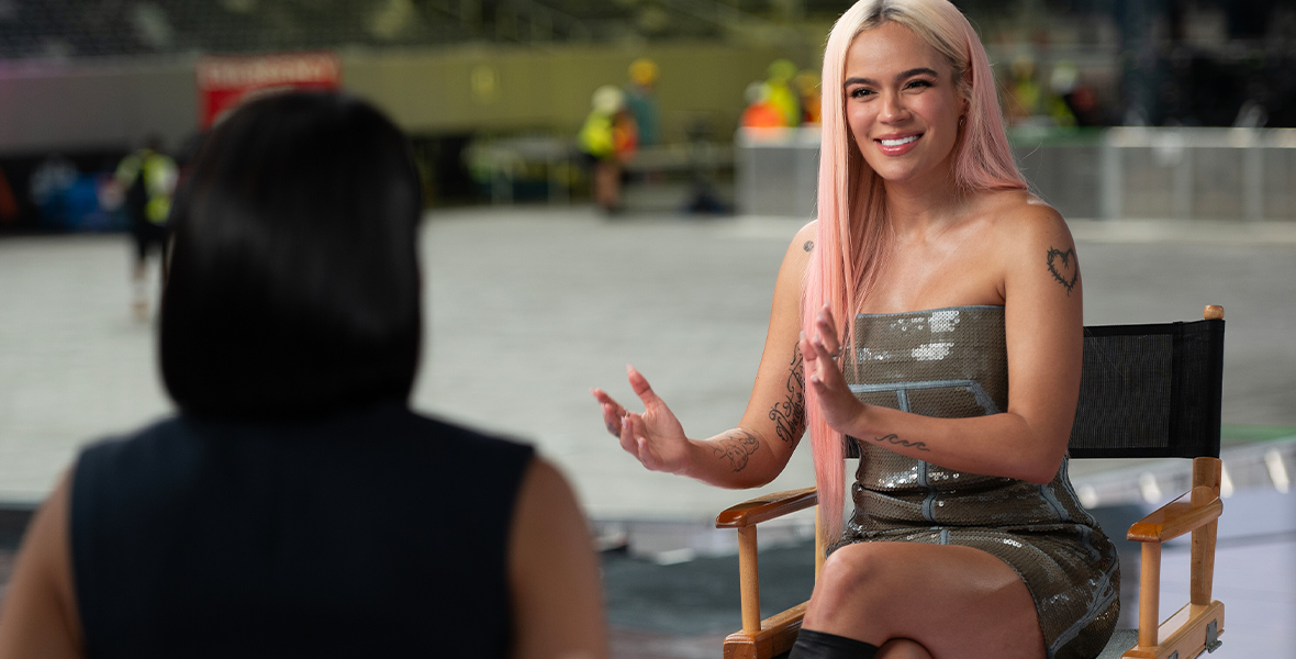 In an image from ABC News’ “The Latin Music Revolution: A Soul of a Nation Presentation,” Colombian artist Karol G is speaking to a reporter, whose back is facing the camera. She’s wearing an army-green sequin sleeveless dress and has long light-pink hair.
