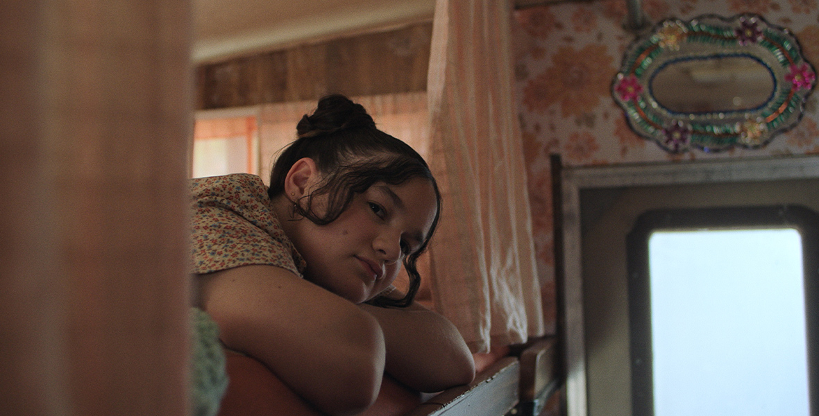 In an image from the Launchpad episode “Beautiful, FL,” a young girl is peering over the side of her bunk bed at something off camera. Her hair is pulled up in a bun, and her head is resting on her folded arms. There are curtains that surround the entrance to the bed, and a door can be seen to the right.