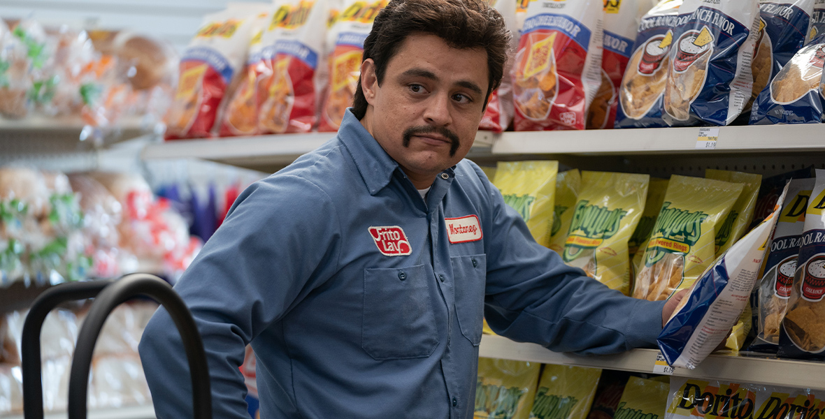 In an image from Hulu’s Flamin’ Hot, Richard Montañez (Jesse Garcia) is standing in front of a large shelving unit full of different Frito Lay chip bags, including Doritos and Funyuns. He’s wearing a blue Frito Lay uniform shirt, that has a nametag patch sewn onto one side of the chest, and a dolly rests on the ground next to him. He has a handlebar mustache.