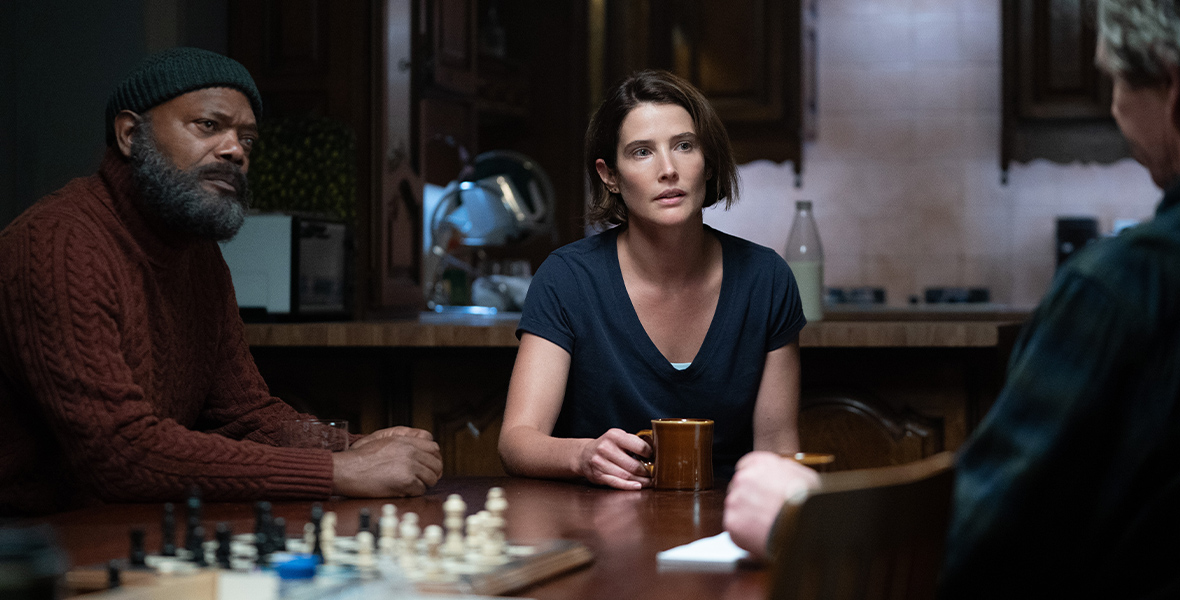 In an image from Marvel Studios’ Secret Invasion, Nick Fury (Samuel L. Jackson) and Maria Hill (Cobie Smulders) are sitting at a table talking with someone who’s just offscreen to the right. There is a chess set sitting on the table. Nick is wearing a burgundy turtleneck sweater and a beanie. Maria is wearing a dark blue V-neck shirt, and is holding a mug in one hand.
