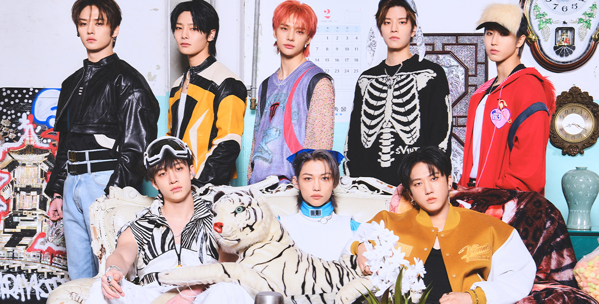 In a promotional image for the 2023 Global Citizen Festival, the popular K-pop group Stray Kids is seen, posed in a living room-like set. Several of the members are sitting on a couch, with a stuffed lion lying across their laps. The rest of the members are standing behind the couch. All are wearing colorful clothing. There is a coffee table in front of the couch, with several cups and a thermos, and a vase of flowers. 