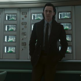 Loki (Tom Hiddleston) stands in front of a bunch of see-through compartments with slices of green pie, topped with whipped cream with his hands in his pocket. He wears a black blazer, black tie, collared shirt, and black plants.