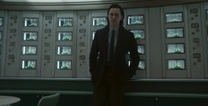 Loki (Tom Hiddleston) stands in front of a bunch of see-through compartments with slices of green pie, topped with whipped cream with his hands in his pocket. He wears a black blazer, black tie, collared shirt, and black plants.