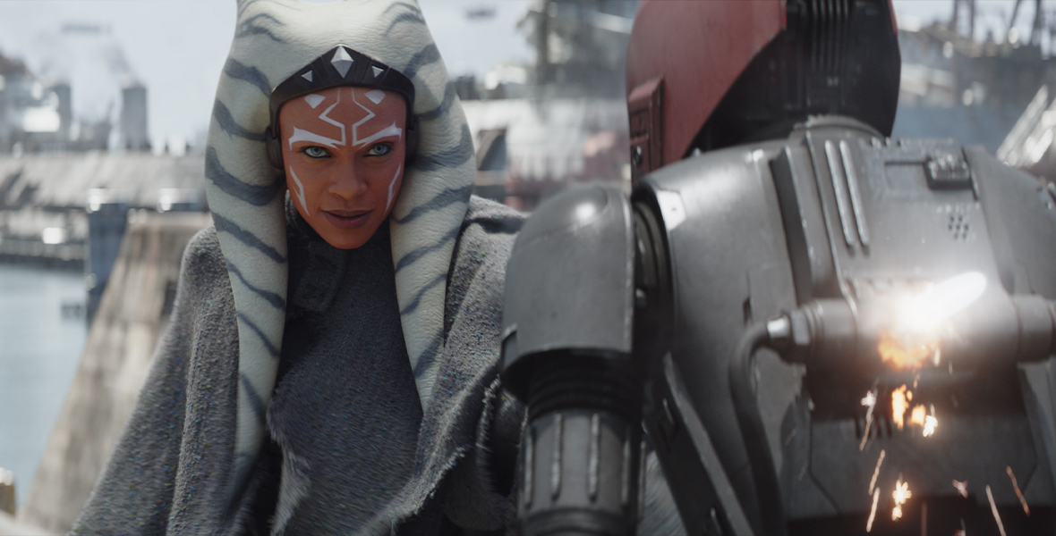 Ahsoka Tano (Rosario Dawson) wears a light gray cloak and stares at a robot with a maroon head and gray body.