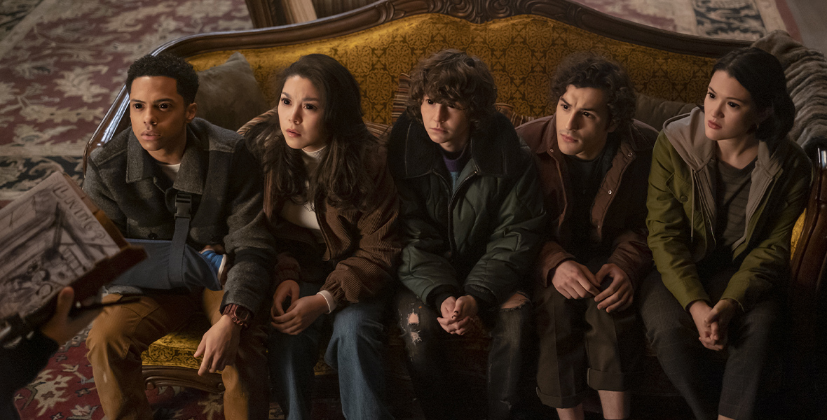 The five kids from the show Goosebumps sit on a yellow couch with a wooden frame while looking up at someone holding a book in their hands with their black eyes. From left to right, Isaiah played by Zack Morris is wearing a gray coat and brown pants, Isabella played by Ana Yi Puig is wearing a brown coat and blue jeans, James played by Miles McKenna is wearing a green puffer coat and ripped black jeans, Edward played by Will Price is wearing a brown jacket and black jeans, Margot played by Isa Briones is wearing a green jacket with a gray hoodie and black jeans.