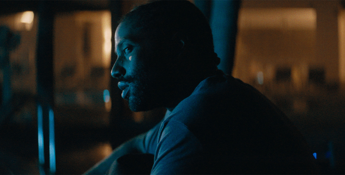 In a scene from the 20th Century Studios film The Creator, John David Washington portrays Joshua, seen here from his left side. He’s wearing a T-shirt and looking to his left with a serious expression on his face. Behind him is a dimly lit and out-of-focus room.