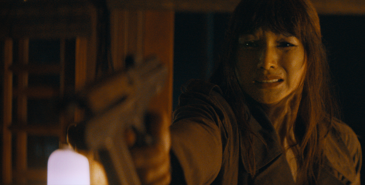 In a scene from the 20th Century Studios film The Creator, Gemma Chan is Maya, seen from the shoulders up. She is wearing a dark tunic and has long, dark hair. She is holding a gun in front of her, close to the camera lens and out of focus, pointed at something out of frame. The room around her is dimly lit but she appears to be standing in a doorway.