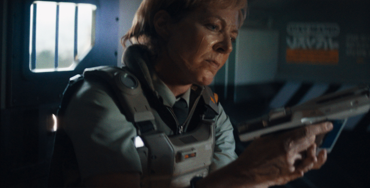 In a scene from the 20th Century Studios film The Creator, Allison Janney is Colonel Jean Howell, seen from her chest up as she loads a handgun in front of her. She is wearing a light teal military jumpsuit with a protective vest over it and appears to be inside some kind of troop transport vehicle, although all that can be seen behind her are two out of focus windows.