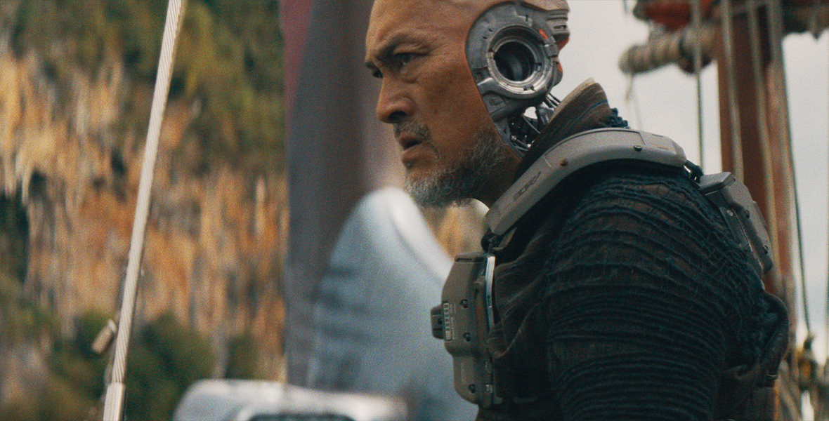 In a scene from the 20th Century Studios film The Creator, the AI warrior Harun is seen from the left side, from the chest up. He is played by Ken Watanabe and is wearing a ribbed black shirt with long sleeves and some kind of weapons vest over it. Instead of ears, he has a metallic cylinder that goes from one side of his head to the other, and the lower back part of his head is entirely robotic. There are indistinct vertical cables and ropes around him and an out of focus mountainside in the distance behind him.