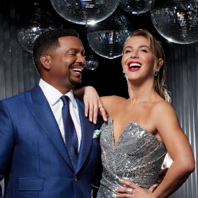 Dancing with the Stars co-host Alfonso Ribeiro (left) wears a blue suit and tie and smiles at co-host Julianne Hough, who wears a strapless silver dress and has her right arm draped on his shoulder. More than a dozen shimmering disco balls hang above their heads.