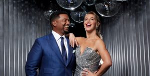 Dancing with the Stars co-host Alfonso Ribeiro (left) wears a blue suit and tie and smiles at co-host Julianne Hough, who wears a strapless silver dress and has her right arm draped on his shoulder. More than a dozen shimmering disco balls hang above their heads.