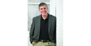 Bestselling young adult novelist Rick Riordan, stands smiling at the camera. He wears a black polo shirt beneath a grey tweed blazer and khaki pants.