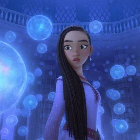 Asha (voiced by Ariana DeBose) is standing in King Magnifico’s observatory, surrounded by floating transparent globes that represent the wishes of the people of Rosas. She looks concerned and is wearing a long-sleeved lavender dress. Behind her are the columns and windows of the 12-sided room, with a balcony visible above her head, running around the inside of the dome above her.