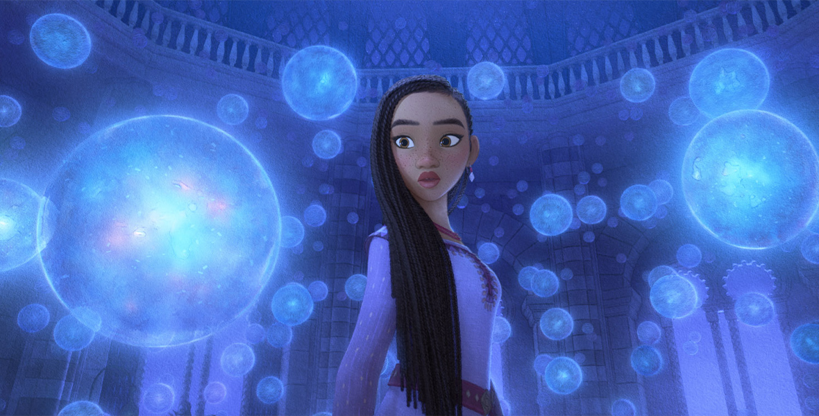 Asha (voiced by Ariana DeBose) is standing in King Magnifico’s observatory, surrounded by floating transparent globes that represent the wishes of the people of Rosas. She looks concerned and is wearing a long-sleeved lavender dress. Behind her are the columns and windows of the 12-sided room, with a balcony visible above her head, running around the inside of the dome above her.