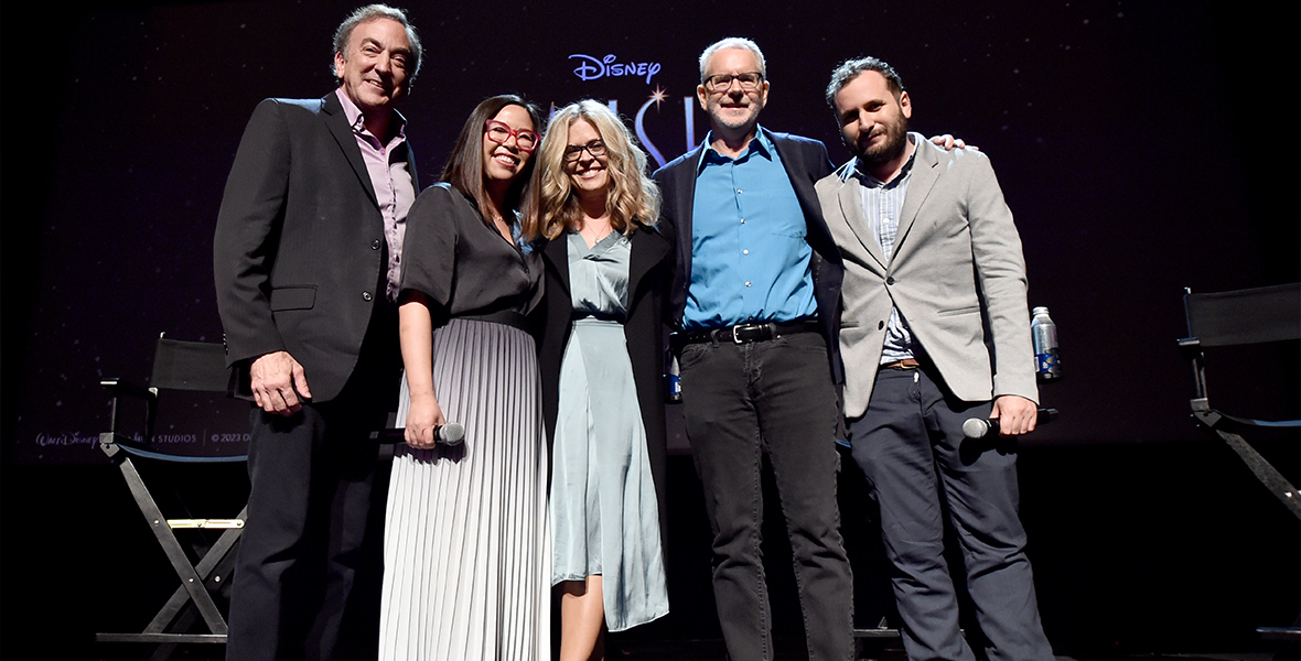 Standing on the stage at the El Capitan Theatre in Hollywood, in a line, are the top creative team from the Walt Disney Animation Studios feature Wish. They are (from left to right) producer Peter Del Vecho, director Fern Veerasunthorn, Walt Disney Animation Studios Chief Creative Officer and screenwriter Jennifer Lee, director Chris Buck, and producer Juan Pablo Reyes. Behind them are directors’ chairs and, barely visible, the logo for the film Wish.