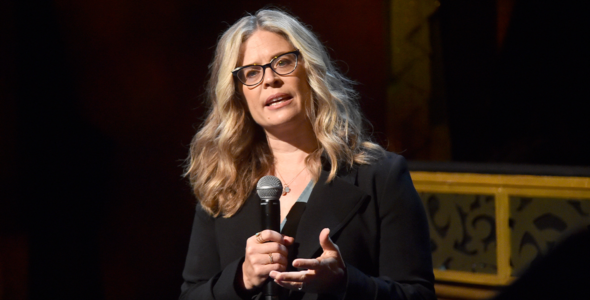 Walt Disney Animation Studios Chief Creative Officer Jennifer Lee stands with a microphone at the El Capitan Theatre in Hollywood, talking about the upcoming feature film Wish. She is wearing glasses and a black suit coat jacket over a blue dress. The room is dark and a bit of the edge of the stage is visible behind her.