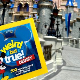A copy of Weird But True! Disney is held up in front of Sleeping Beauty Castle at Magic Kingdom Park. The cover of the book features the title superimposed over a blue, three-circle Mickey. To the left of the title, a classic version of Mickey Mouse points to himself with a hand on his hip.