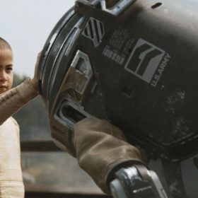 In a scene from the film The Creator, Alphie, played by Madeleine Yuna Voyles, is wearing a beige long-sleeve shirt with a red patch on her right shoulder while she puts her right hand on a metal canister-shaped robot that has 4 metal legs.