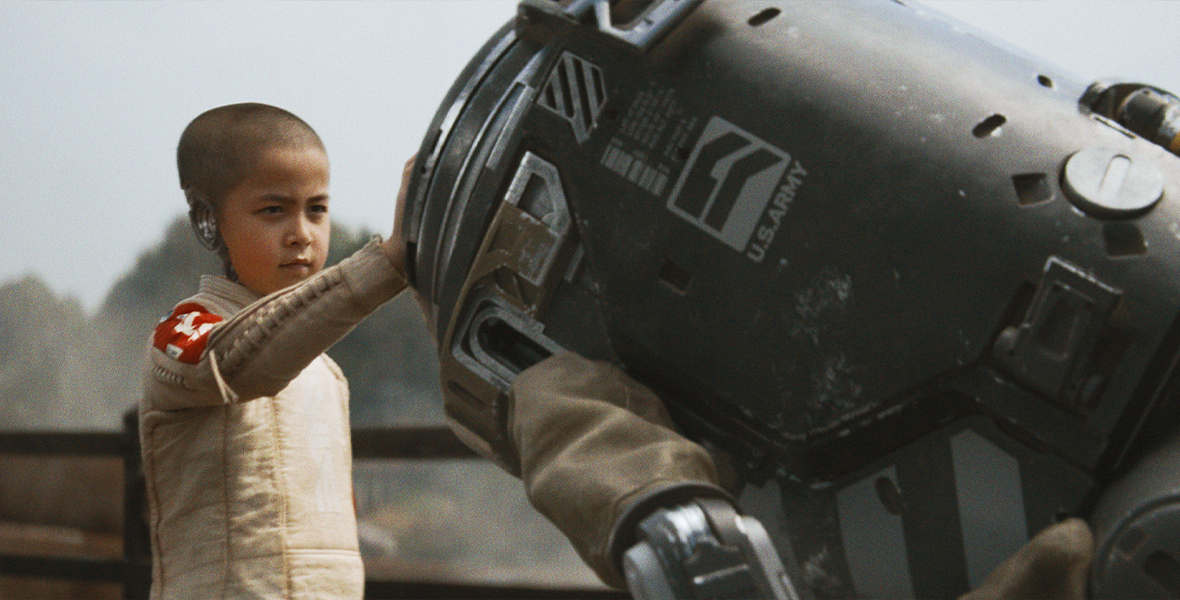 In a scene from the film The Creator, Alphie, played by Madeleine Yuna Voyles, is wearing a beige long-sleeve shirt with a red patch on her right shoulder while she puts her right hand on a metal canister-shaped robot that has 4 metal legs.