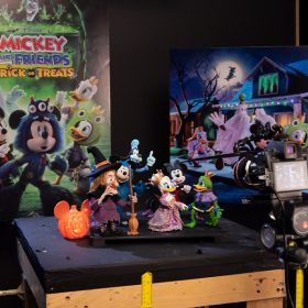 On a wooden worktable topped with black fabric sits a group of the stop-motion puppets from the Halloween special Mickey and Friends Trick or Treats. The puppets include Witch Hazel and five Disney characters in Halloween costumes: Mickey Mouse as a blue monster, Minnie Mouse as a spider, Daisy Duck as a princess, Donald Duck as a frog prince, and Goofy as a ghost. Behind the puppet display is a poster for the show and an enlarged print of a still image from the special, depicting the Disney characters trick-or-treating on a street.