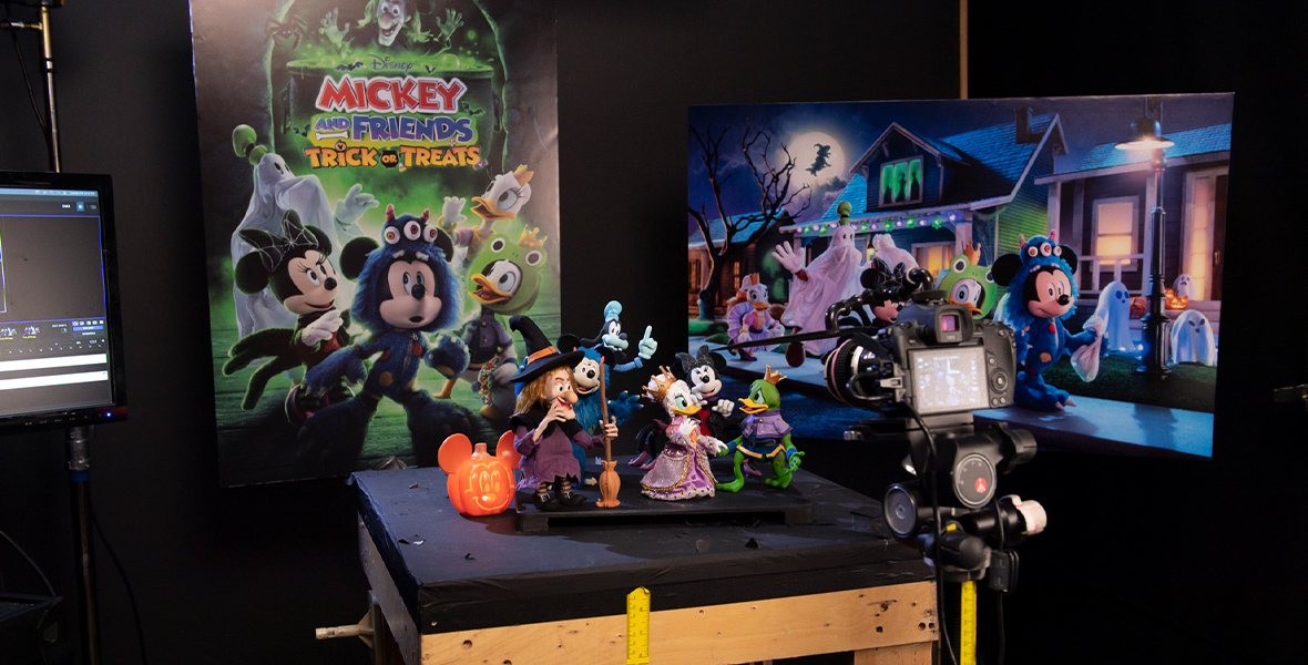 On a wooden worktable topped with black fabric sits a group of the stop-motion puppets from the Halloween special Mickey and Friends Trick or Treats. The puppets include Witch Hazel and five Disney characters in Halloween costumes: Mickey Mouse as a blue monster, Minnie Mouse as a spider, Daisy Duck as a princess, Donald Duck as a frog prince, and Goofy as a ghost. Behind the puppet display is a poster for the show and an enlarged print of a still image from the special, depicting the Disney characters trick-or-treating on a street.
