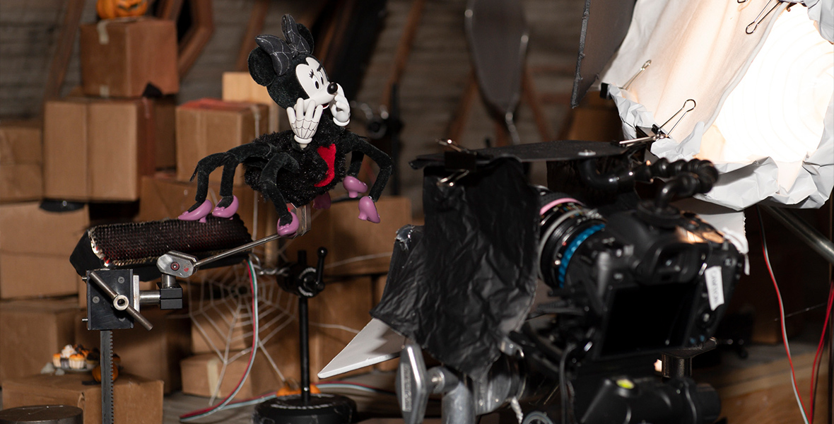 The stop-motion puppet representing Minnie Mouse, in the form of the six-legged Spider Minnie, is suspended in the air in front of an elaborate camera setup. Behind her is the miniature set for Witch Hazel’s attic.