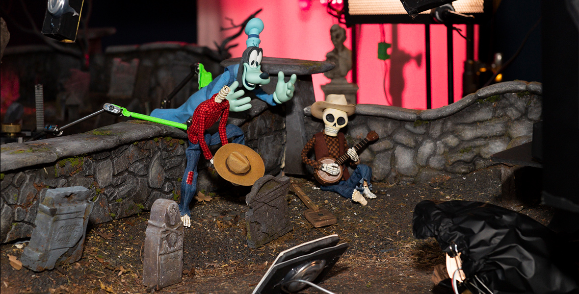 In a close-up image of a stop-motion set, Ghost Goofy is seen hovering over a wall in a graveyard. He has his hands raised in an effort to frighten the two skeleton men on either side of him. The skeletons are both dressed in shirts and jeans. The skeleton on the right is leaning against the cemetery wall, playing a banjo, while the skeleton on the left has jumped up so quickly that his head has come off. The missing skull is nowhere to be seen.