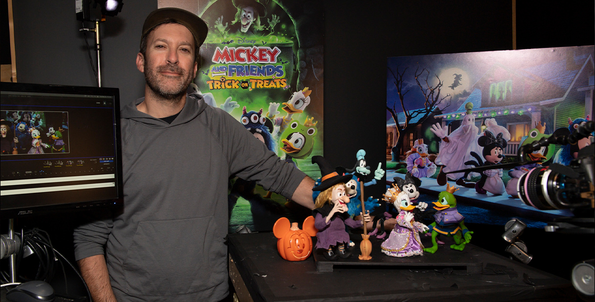 David Brooks, the director and producer of Mickey and Friends Trick or Treat, stands next to a wooden worktable topped with black fabric. On the table sits a group of the stop-motion puppets from the Halloween special. The puppets include Witch Hazel and five Disney characters in Halloween costumes: Mickey Mouse as a blue monster, Minnie Mouse as a spider, Daisy Duck as a princess, Donald Duck as a frog prince, and Goofy as a ghost. Behind the puppet display is a poster for the show and an enlarged print of a still image from the special, depicting the Disney characters trick-or-treating on a street.