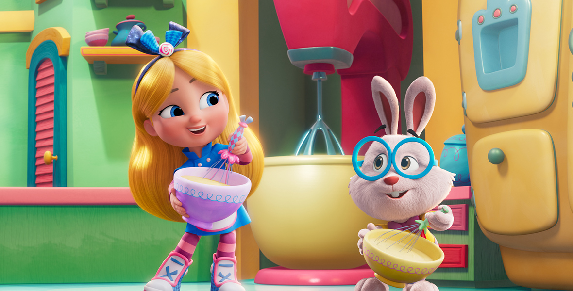 In an image from Disney Branded Television’s Alice’s Wonderland Bakery, Alice (voiced by Libby Rue), left, and Fergie (voiced by Jack Stanton), right, are standing in a kitchen and both holding mixing bowls and whisks. Behind them is a larger-than-life standing mixer, as well as an oven to their right. Fergie, a bunny, is wearing large blue glasses.