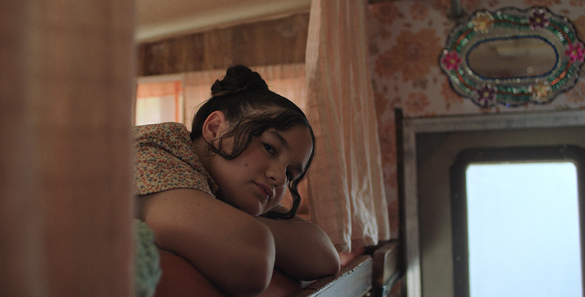 In an image from the Launchpad episode “Beautiful, FL,” a young girl named Omara (Dariana Alvarez) is peering over the side of her bunk bed at something off camera. Her hair is pulled up in a bun, and her head is resting on her folded arms. There are curtains that surround the entrance to the bed, and a door can be seen to the right.