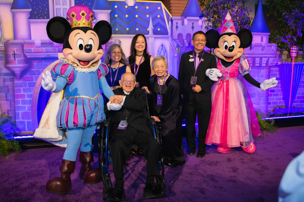 A family pose with Mickey Mouse and Minnie Mouse. Mickey is dressed in a blue and pink outfit with puff sleeves, brown boots, and a pink and gold crown. Minnie is wearing a pink princess outfit with a pink pointy princess hat and pink shoes. A man in a wheel chair holds Mickey Mouse’s hand, a woman stands next to his wheel chair while 3 people stand behind the man and woman while smiling.