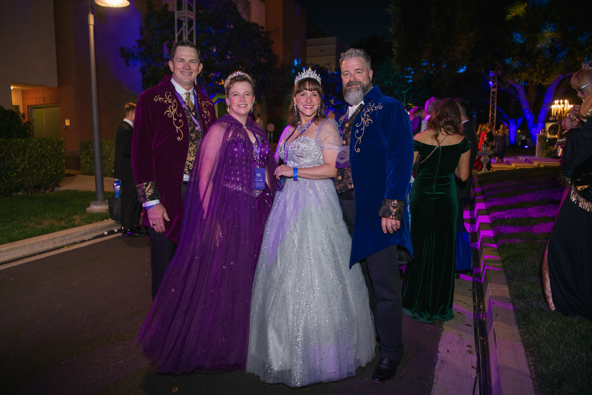 Two couples pose together. From left to right, a man is wearing a long maroon coat with gold detailing and black slacks, next to him a woman wears a purple, sleeveless dress with a sheer purple cape and a gold tiara. Next to her, a woman wears a light blue sparkly dress with sheer puff sleeves and silver tiara. Next to her, a man wears a blue coat with gold detailing and black slacks.