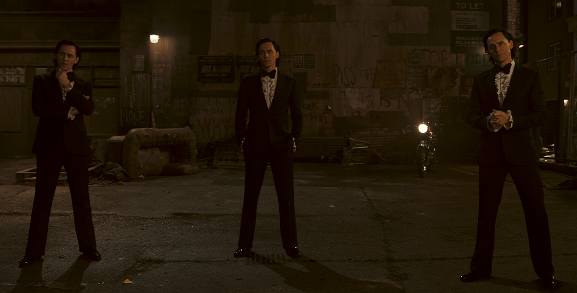 A trio of Lokis, all played by Tom Hiddleston, wear '80s-style tuxedos and ruffled shirts. Each Loki strikes a different pose as they stand in a dimly lit and dirty alley at night.